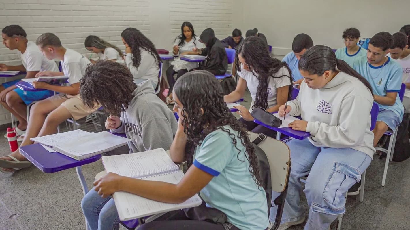 Estudantes sentados em carteiras escrevem em cadernos durante aula em sala de escola pública