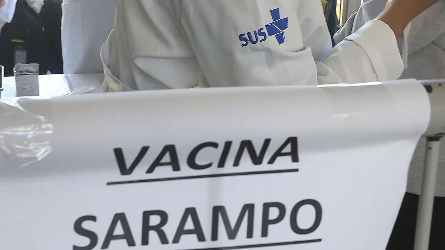Faixa 'Vacina Sarampo' em posto de vacinação com agente de saúde do SUS ao fundo