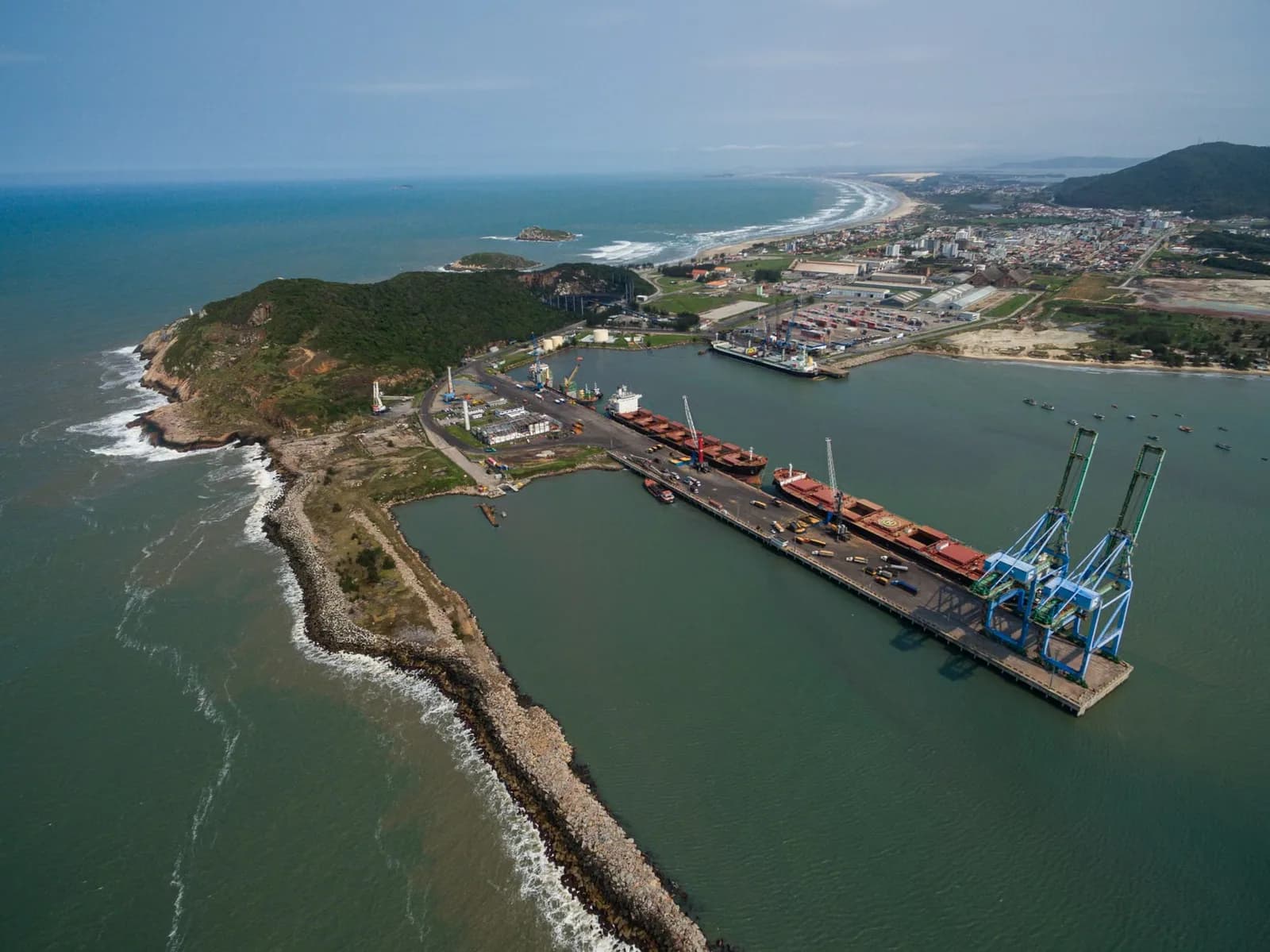 Imagem ilustrativa: Vista aérea de um porto com navios atracados, cercado por mar e uma costa com vegetação e edificações ao fundo