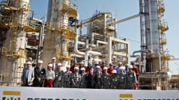 Trabalhadores com capacete reunidos em refinaria da Petrobras, com estruturas industriais e banner da empresa ao fundo