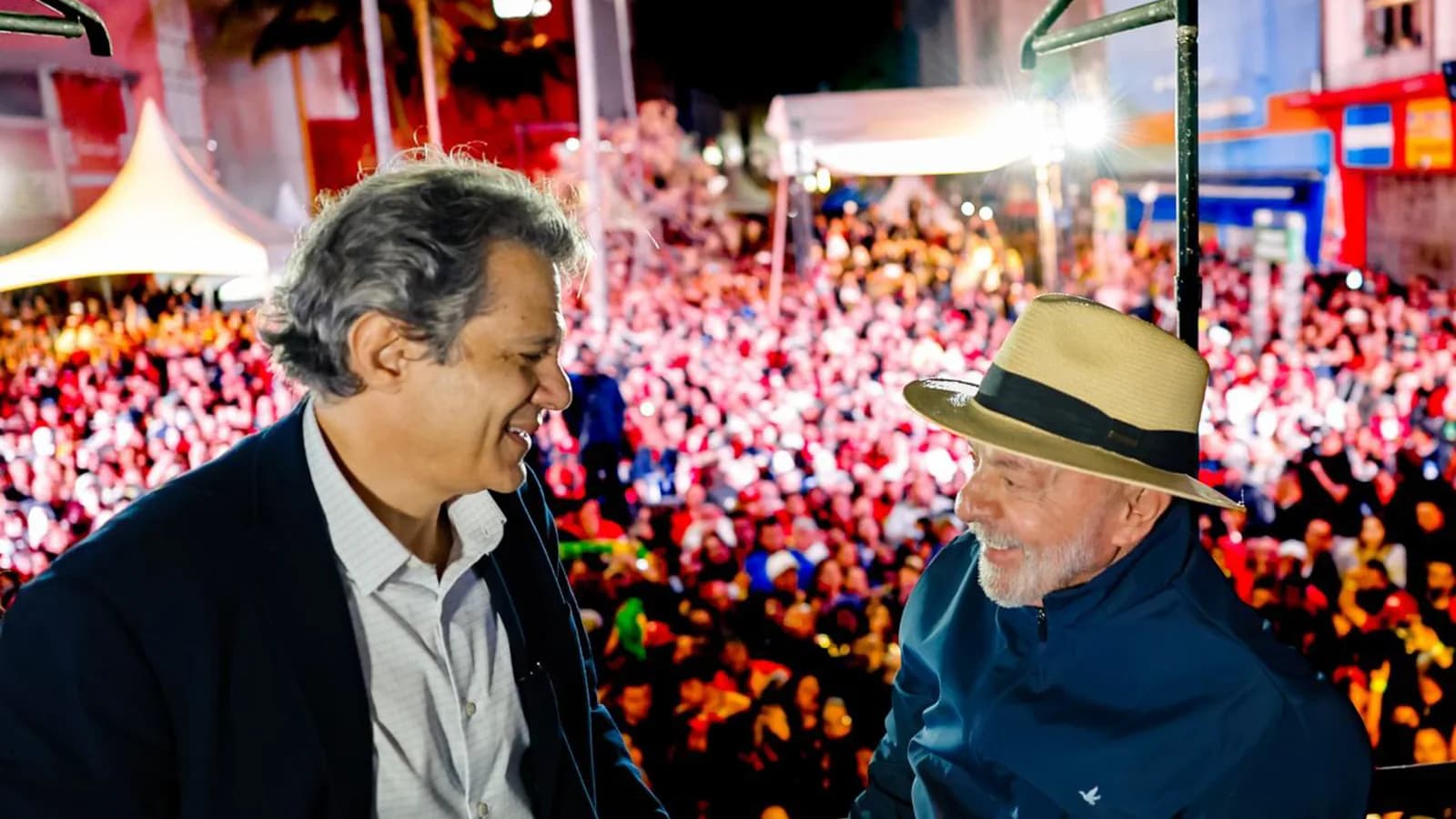 Lula cumprimenta apoiador em comício de campanha à noite, com multidão ao fundo