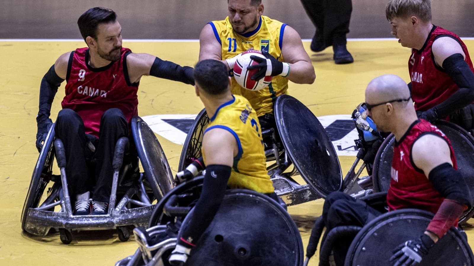 Jogador do Brasil disputa a bola com adversários do Canadá durante partida de rúgbi em cadeira de rodas