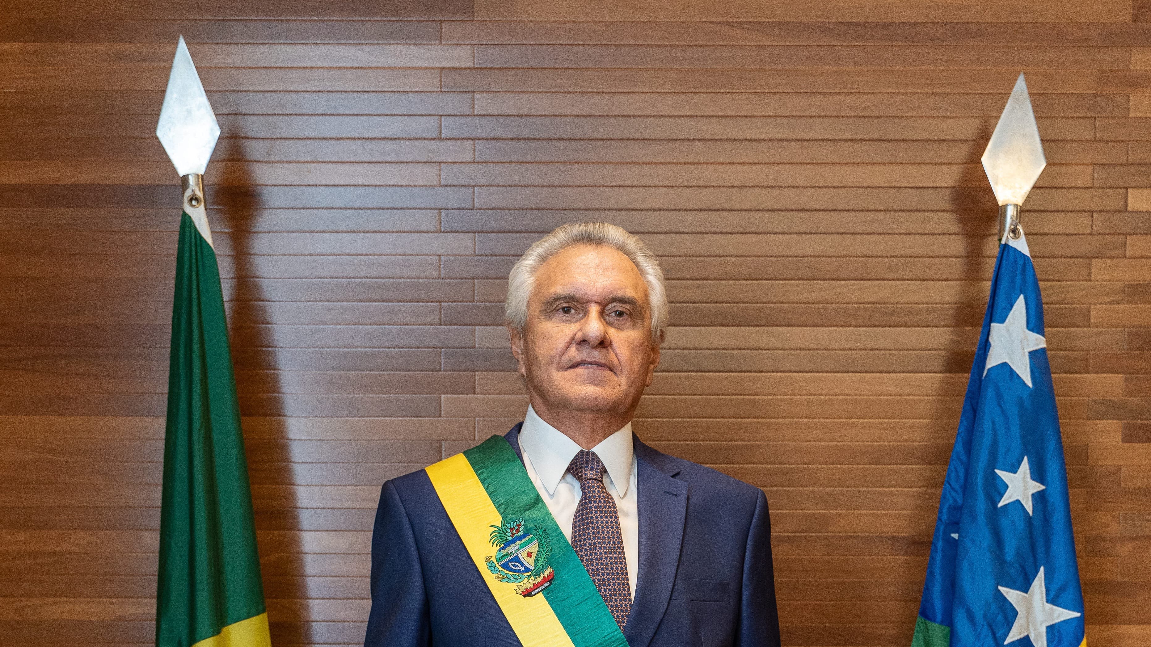 Ronaldo Caiado, governador de Goiás, em foto oficial de posse ao cargo em 2023, com a faixa de governador, ladeado pelas bandeiras do Brasil e de Goiás
