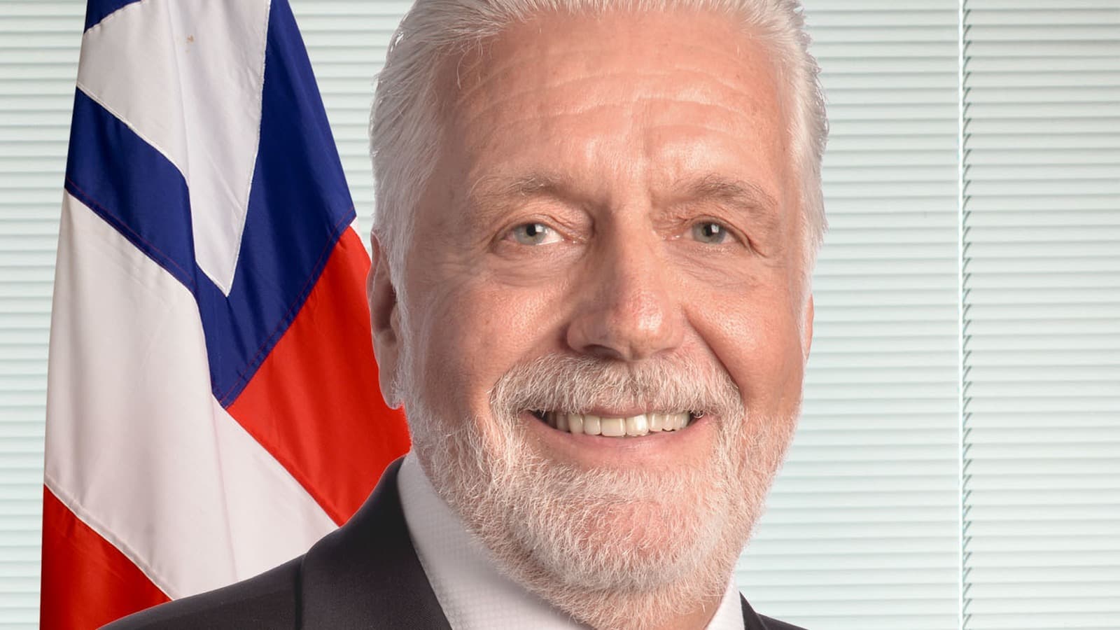Retrato oficial do senador Jaques Wagner, de terno escuro e gravata vermelha, diante da bandeira da Bahia