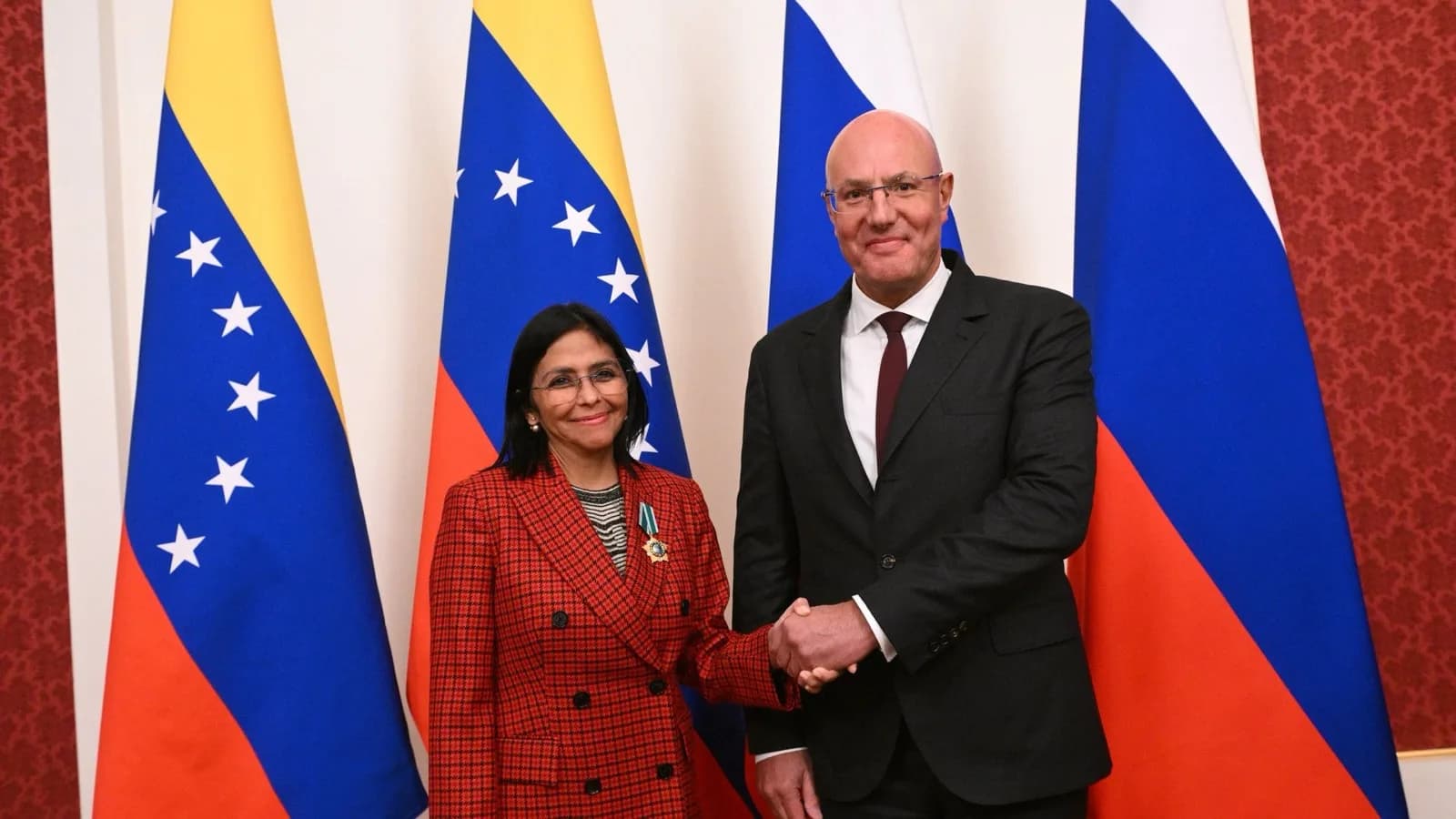 Delcy Rodríguez, presidente interina da Venezuela, cumprimenta autoridade em evento oficial, com bandeiras da Venezuela e da Rússia ao fundo