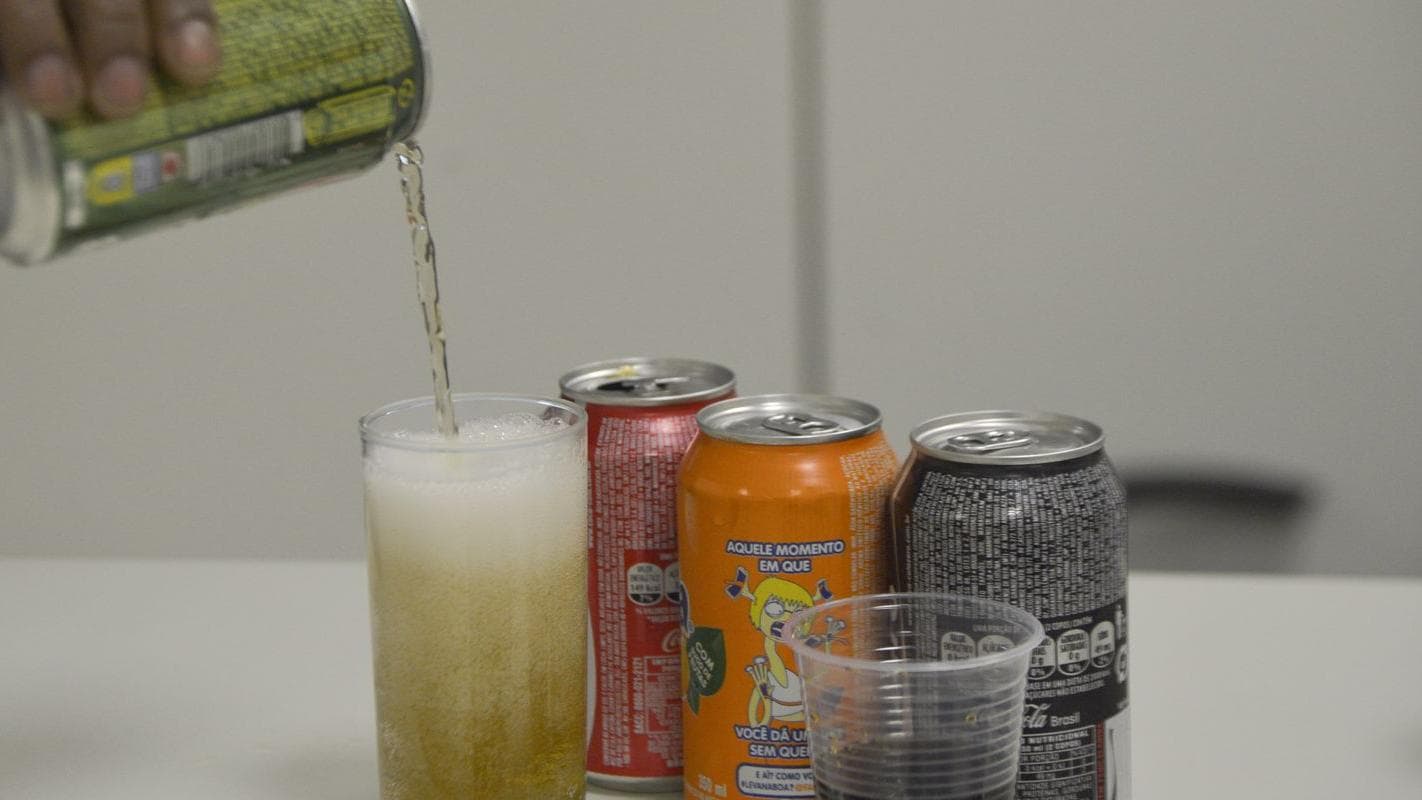 Pessoa serve refrigerante em copo, com outras latas ao fundo, ilustrando consumo de alimentos ultraprocessados