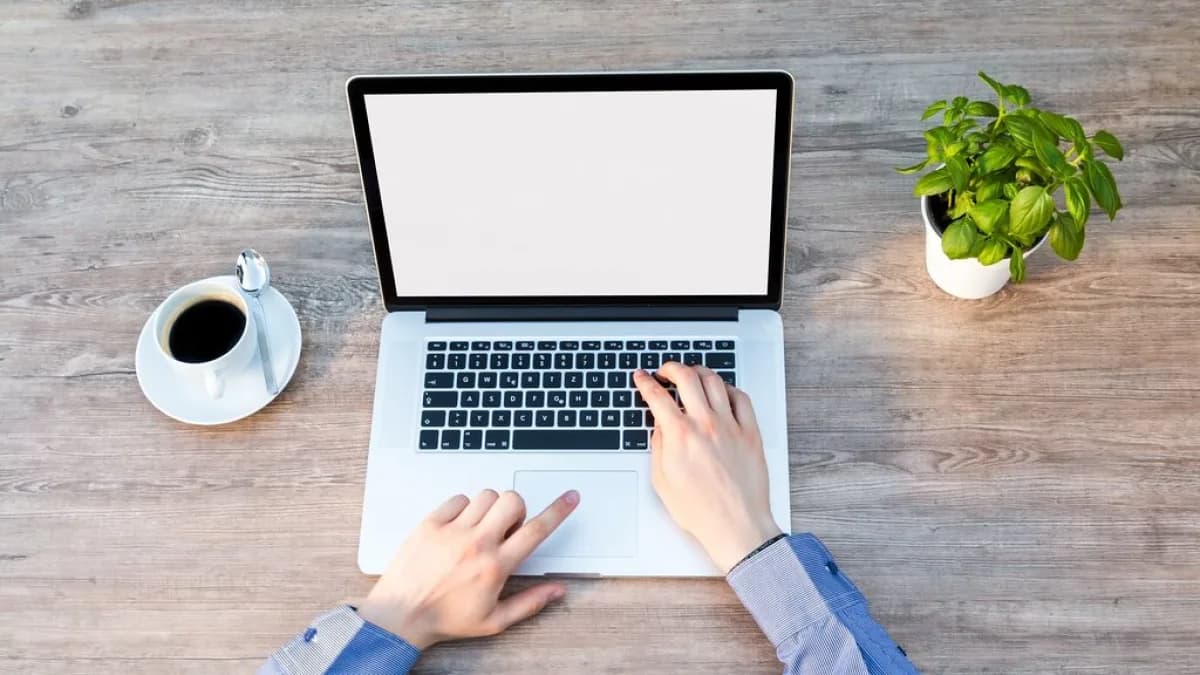 Xícara de café ao lado de um notebook em uma mesa de madeira, com mãos digitando no teclado