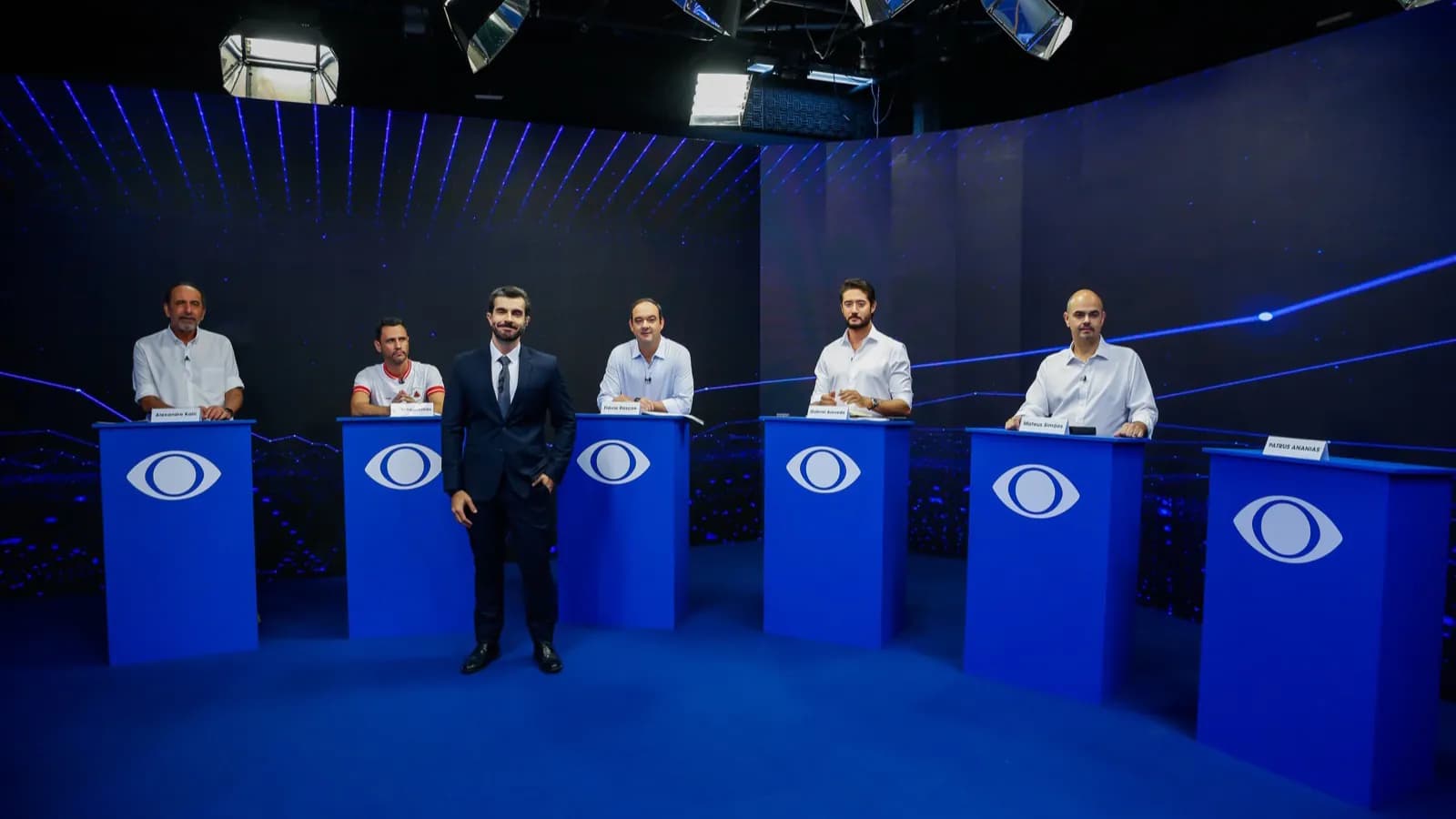 Cinco candidatos ao Governo de Minas Gerais perfilados atrás de púlpitos azuis no estúdio da Band Minas, com o mediador Lucas Catta Prêta ao centro