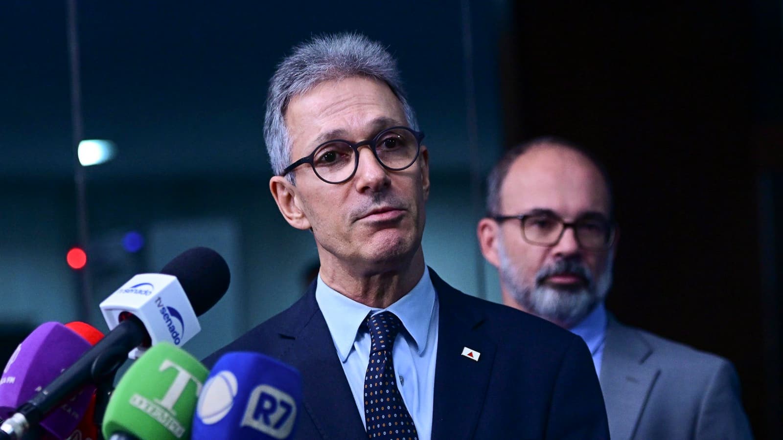 Romeu Zema, de terno escuro, óculos redondos e cabelo grisalho, fala diante de microfones de emissoras de TV em uma entrevista coletiva; ao fundo, desfocado, aparece um homem de terno claro