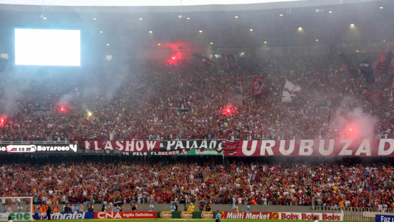 Torcida do Flamengo lotando a arquibancada do Maracanã com fumaça e sinalizadores, faixa Urubuzada estendida