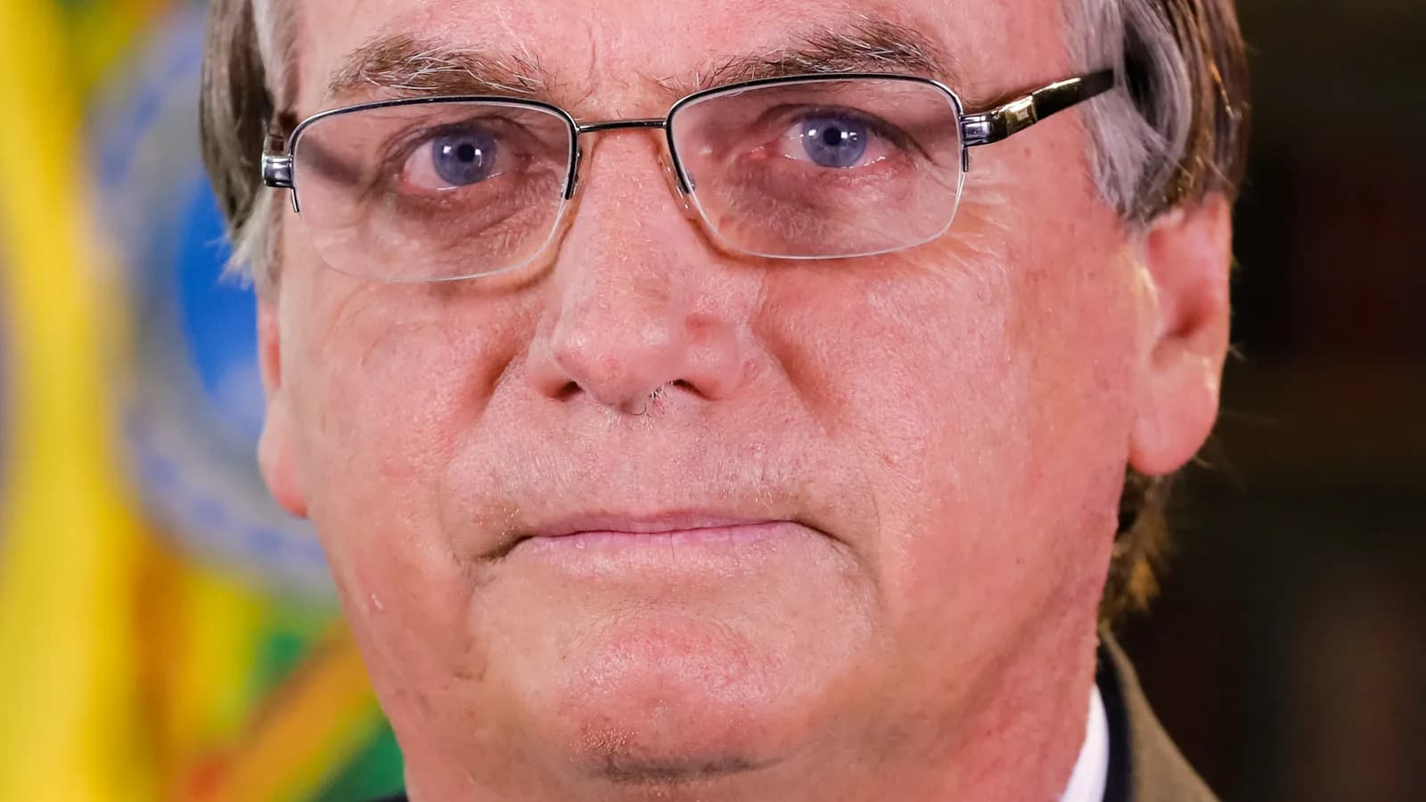 Jair Bolsonaro em retrato sério, de óculos e terno, com a bandeira do Brasil ao fundo