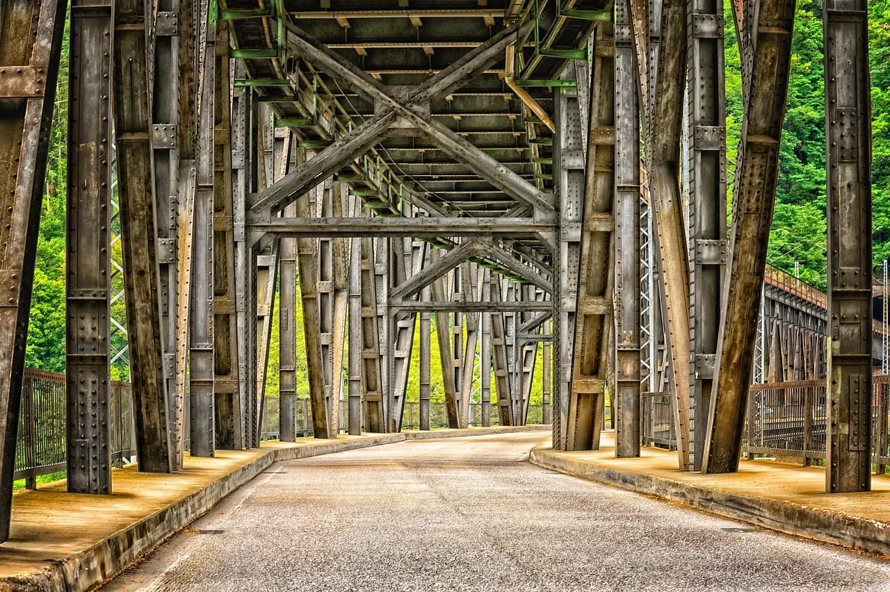 Imagem ilustrativa: Vista interna de um viaduto de metal com estrutura exposta e estrada pavimentada sob um céu claro e vegetação ao fundo