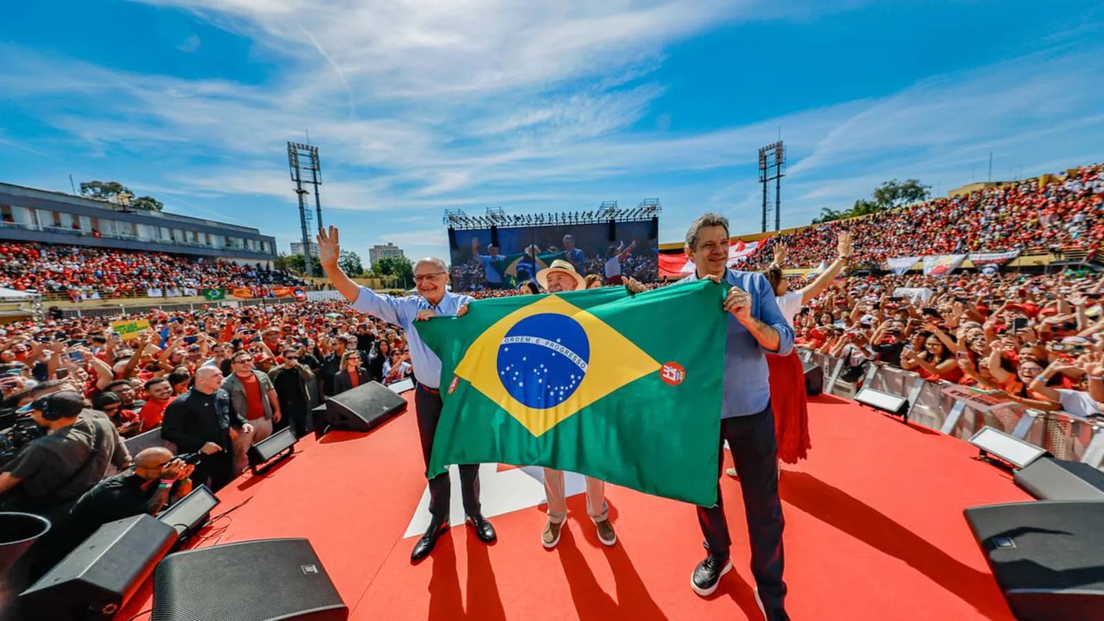 Lula acena para multidão segurando bandeira do Brasil em comício de campanha