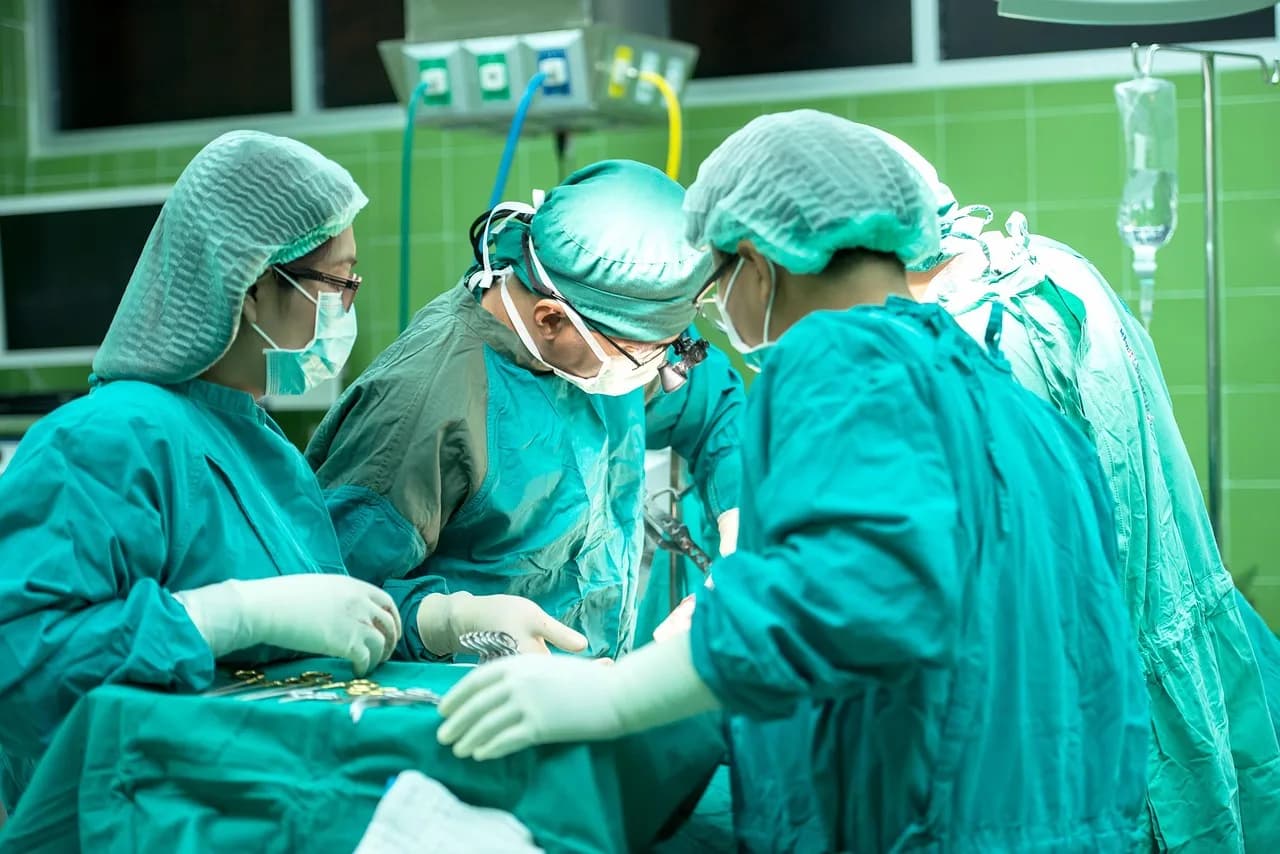 Imagem ilustrativa: Quatro profissionais de saúde em ambiente cirúrgico, usando roupas e equipamentos de proteção, focados em um procedimento.