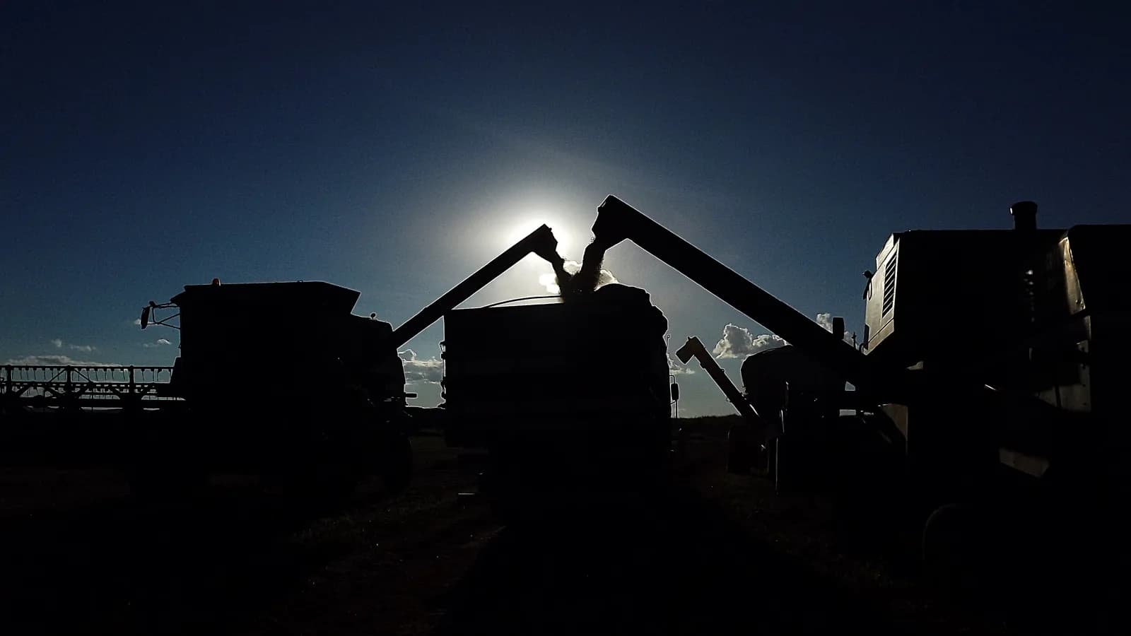 Silhueta de colheitadeiras e caminhões descarregando grãos de soja ao entardecer em fazenda no interior de Goiás