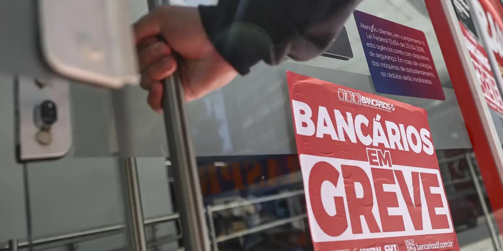 Mão fixa cartaz vermelho com os dizeres Bancários em Greve na porta de uma agência bancária