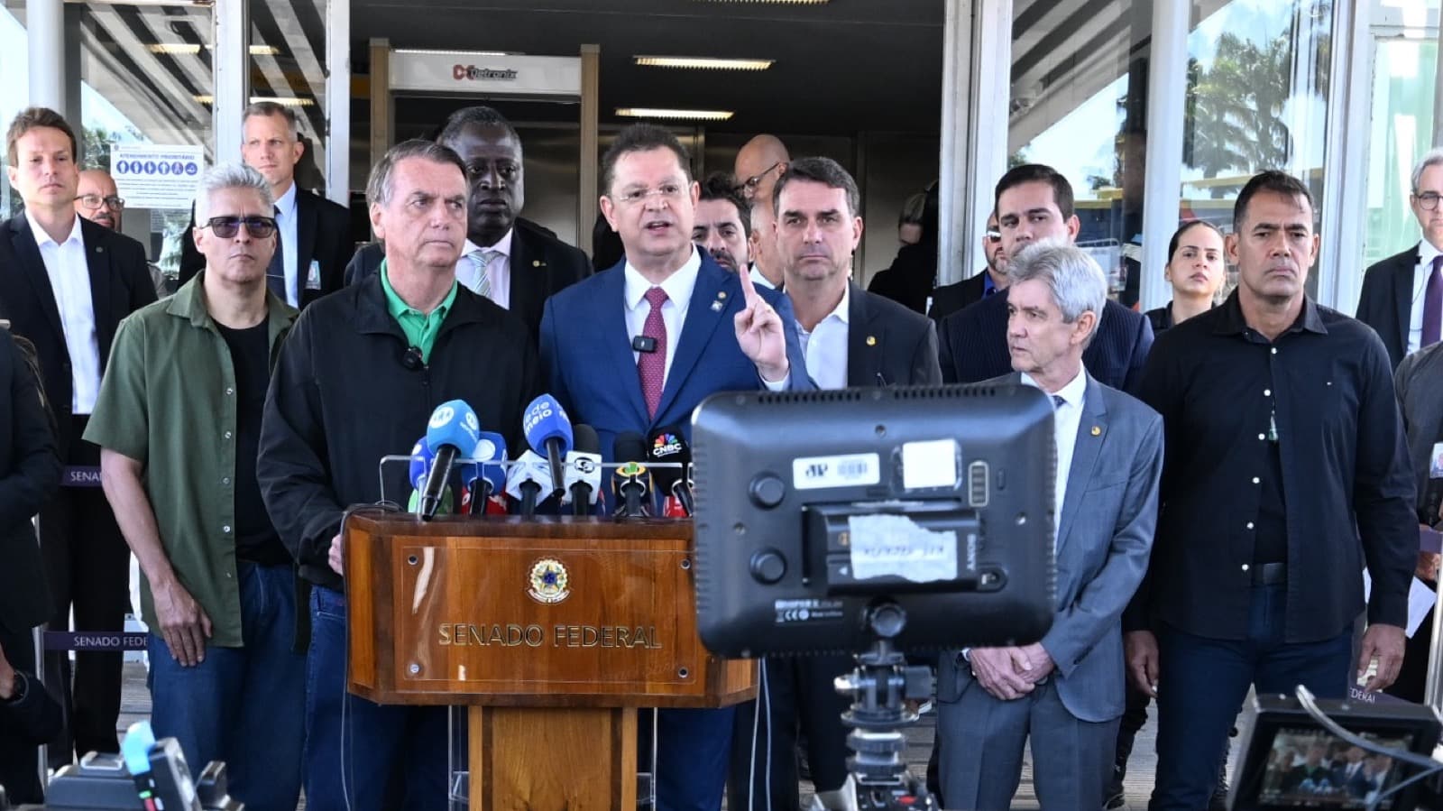 Jair Bolsonaro, de camisa verde, e o senador Flávio Bolsonaro atrás de um púlpito do Senado Federal durante entrevista coletiva, cercados por parlamentares e microfones da imprensa, em imagem de arquivo de julho de 2025