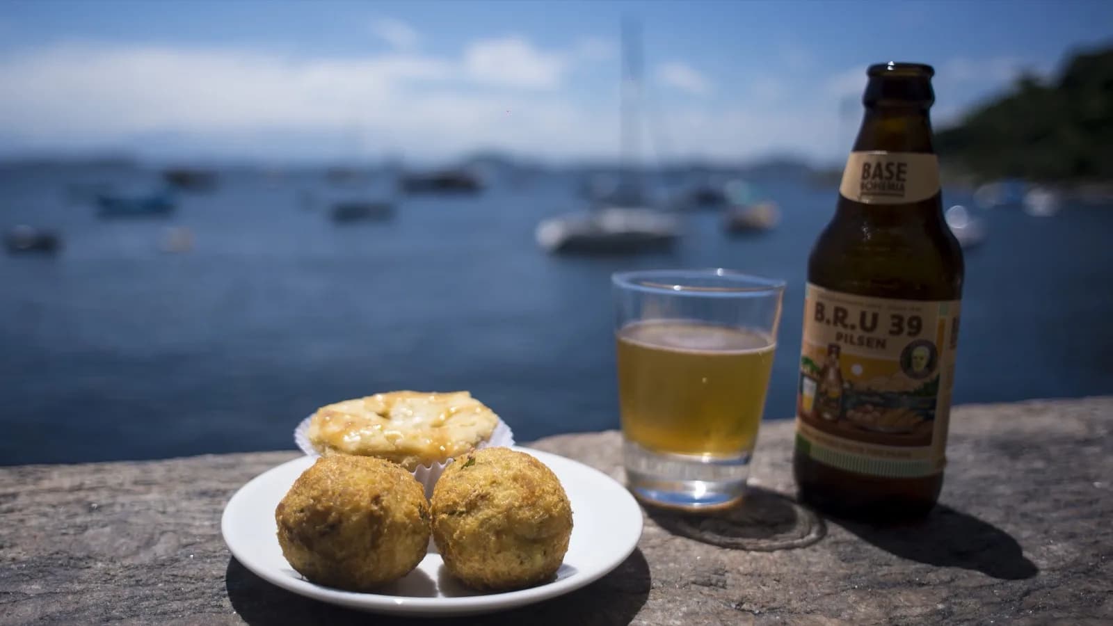 Prato com bolinhos e empada, chope e garrafa de cerveja sobre pedra à beira da baía do Rio de Janeiro, com veleiros ao fundo, foto de arquivo ilustrativa