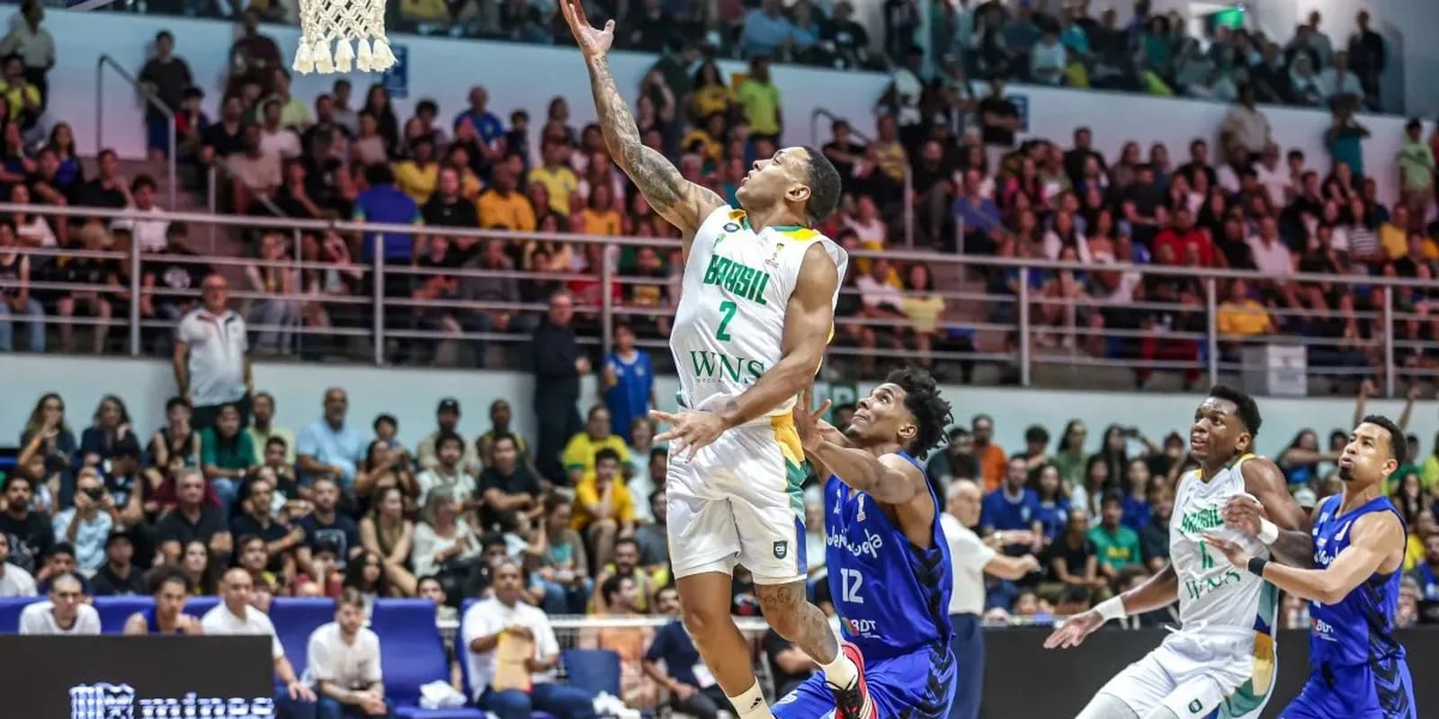 Jogador da seleção brasileira de basquete arremessa ao gol durante partida das Eliminatórias, com defensor da Venezuela tentando bloquear