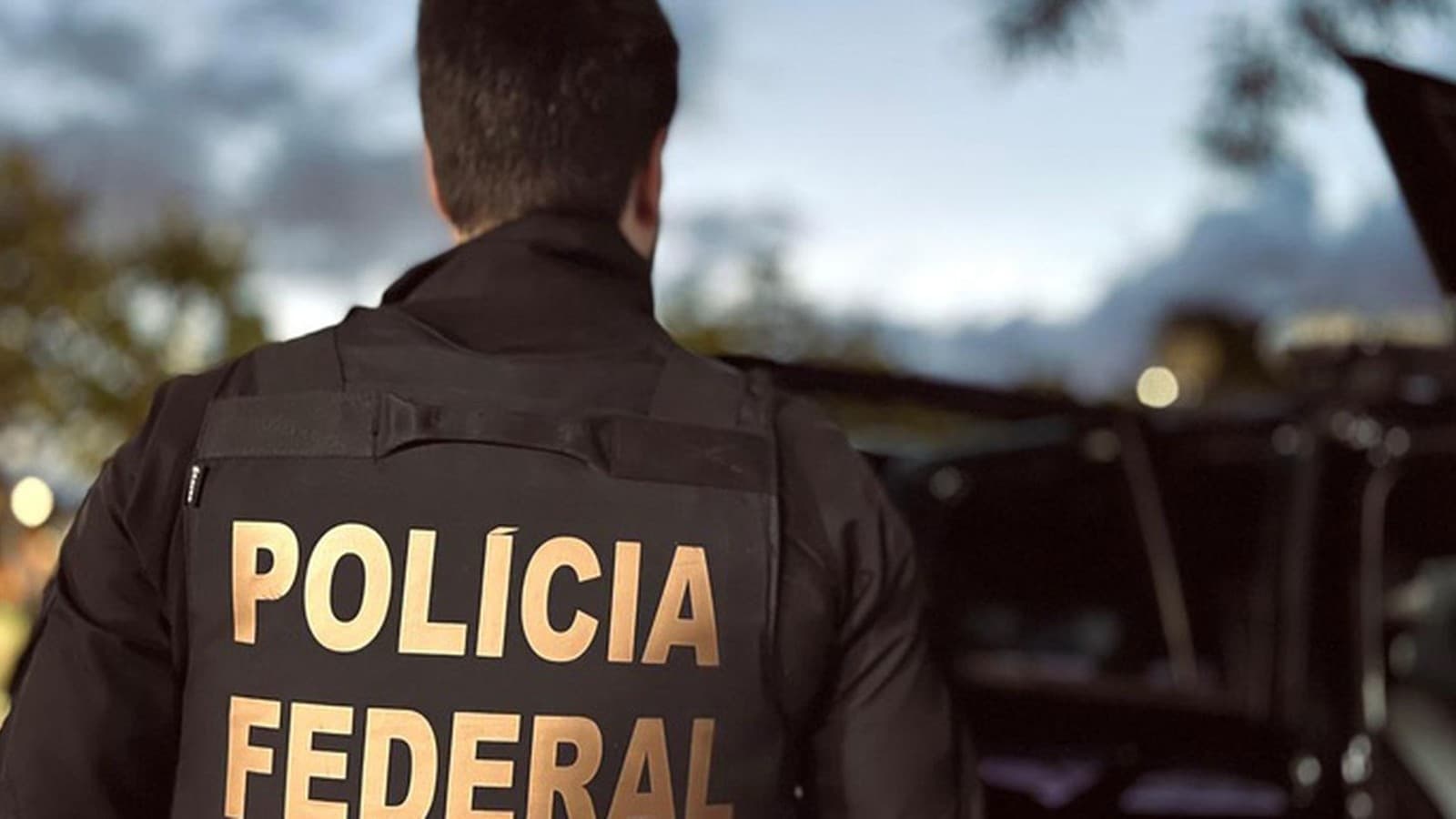 Agente da Polícia Federal visto de costas, com o colete identificado, ao lado de um veículo