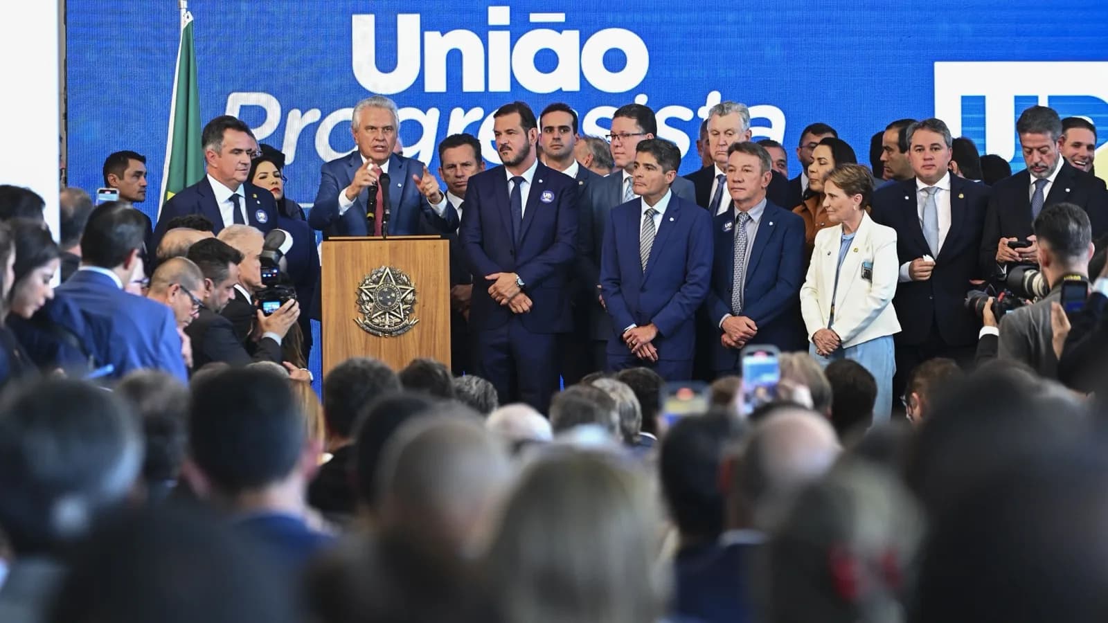 Ronaldo Caiado discursa em púlpito durante evento político, cercado por parlamentares, com telão da União Progressista ao fundo