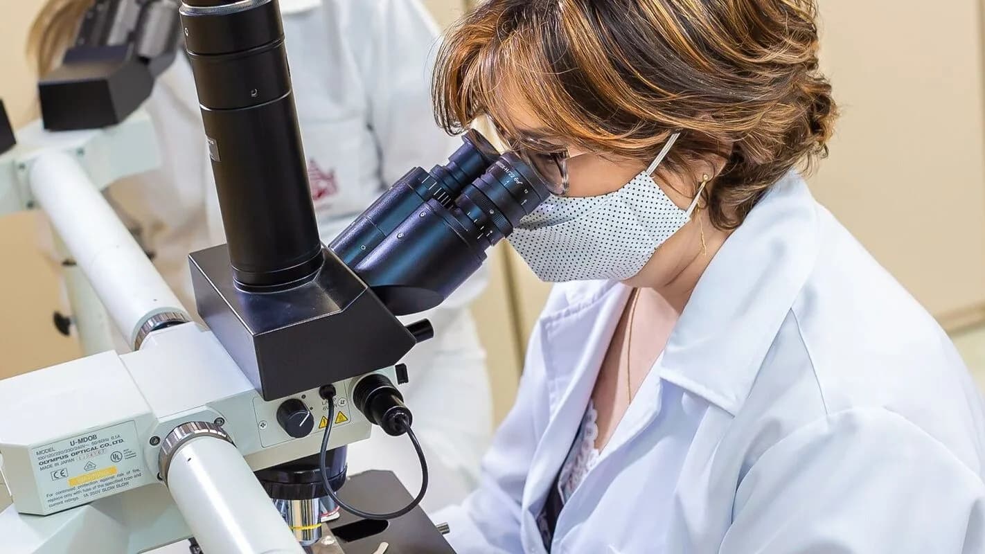 Pesquisadora de jaleco branco observa amostra em microscópio de laboratório